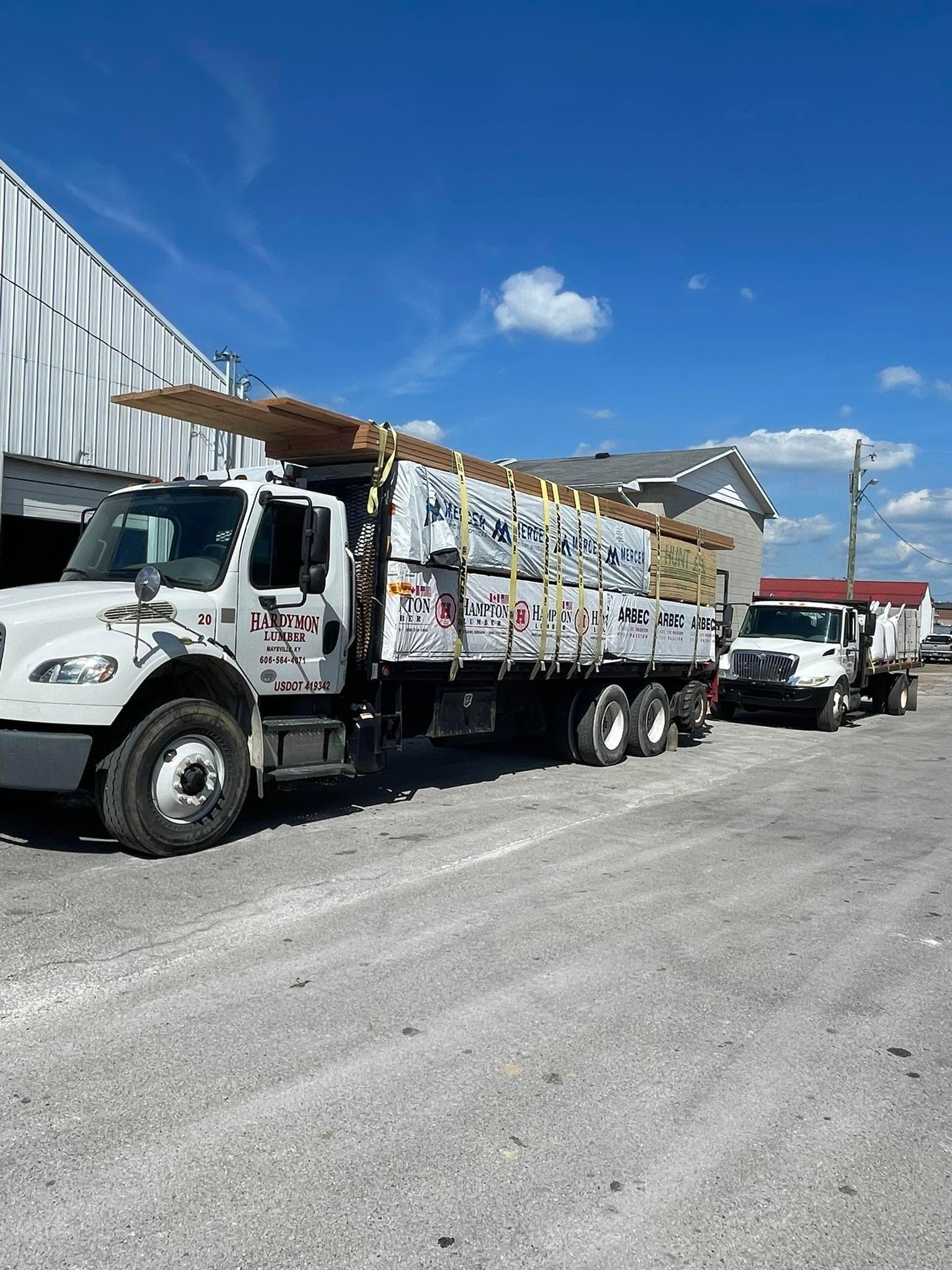 Two white utility trucks parked in an industrial yard beside a corrugated metal building under a blue sky