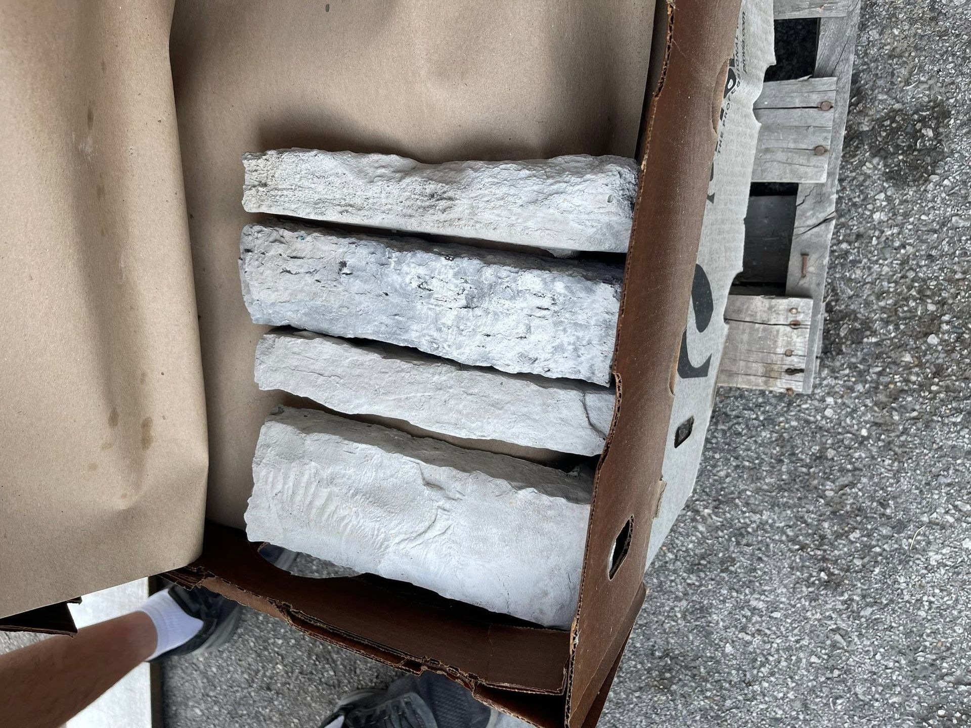 Cardboard box holding stacked white stone or foam slabs on a gravel surface, with a shoe at left.