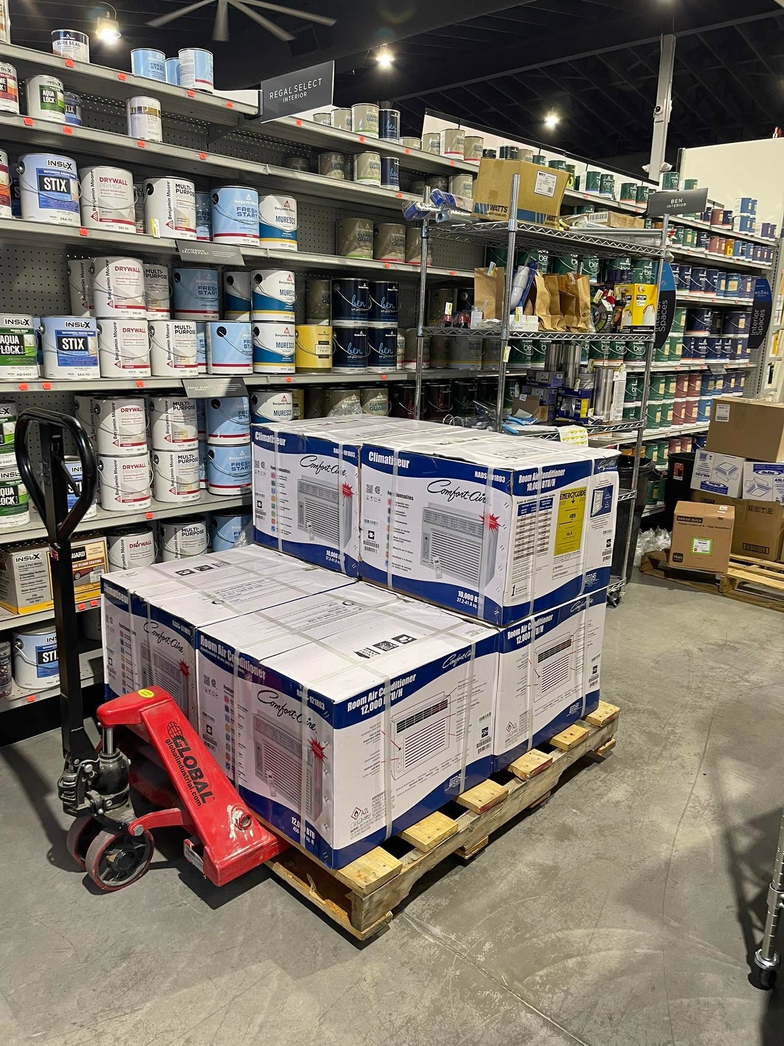 Warehouse aisle with stacked boxes on pallets, shelves of paint buckets, and a red pallet jack in front