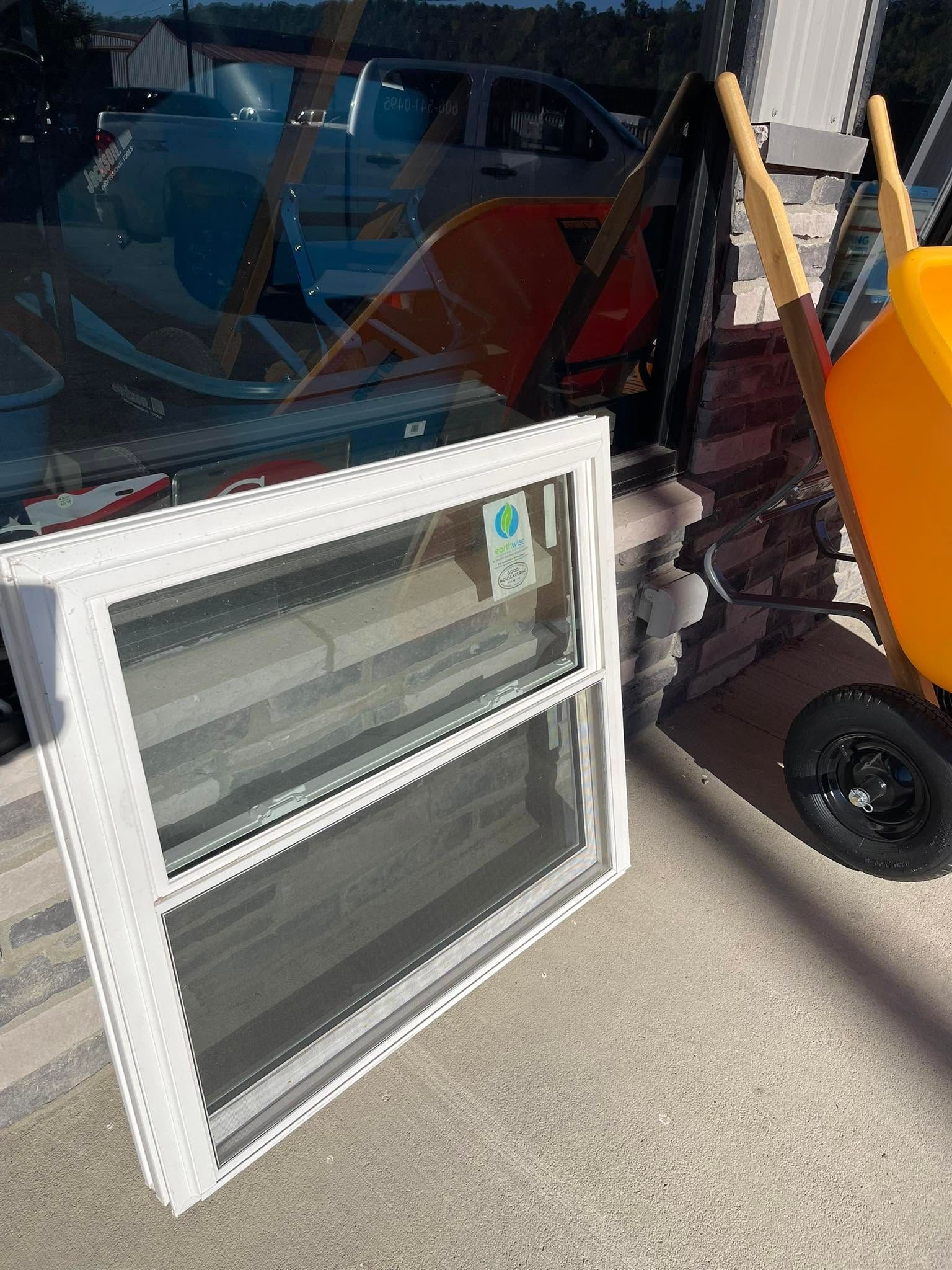 White window leaning against storefront beside orange wheelbarrow and black hand truck