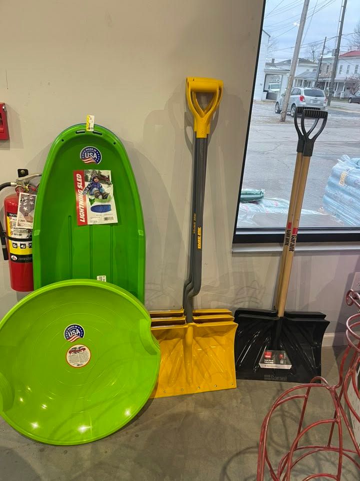 Green sleds and snow shovels leaning against a wall in a store, with a window showing snowy street outside.