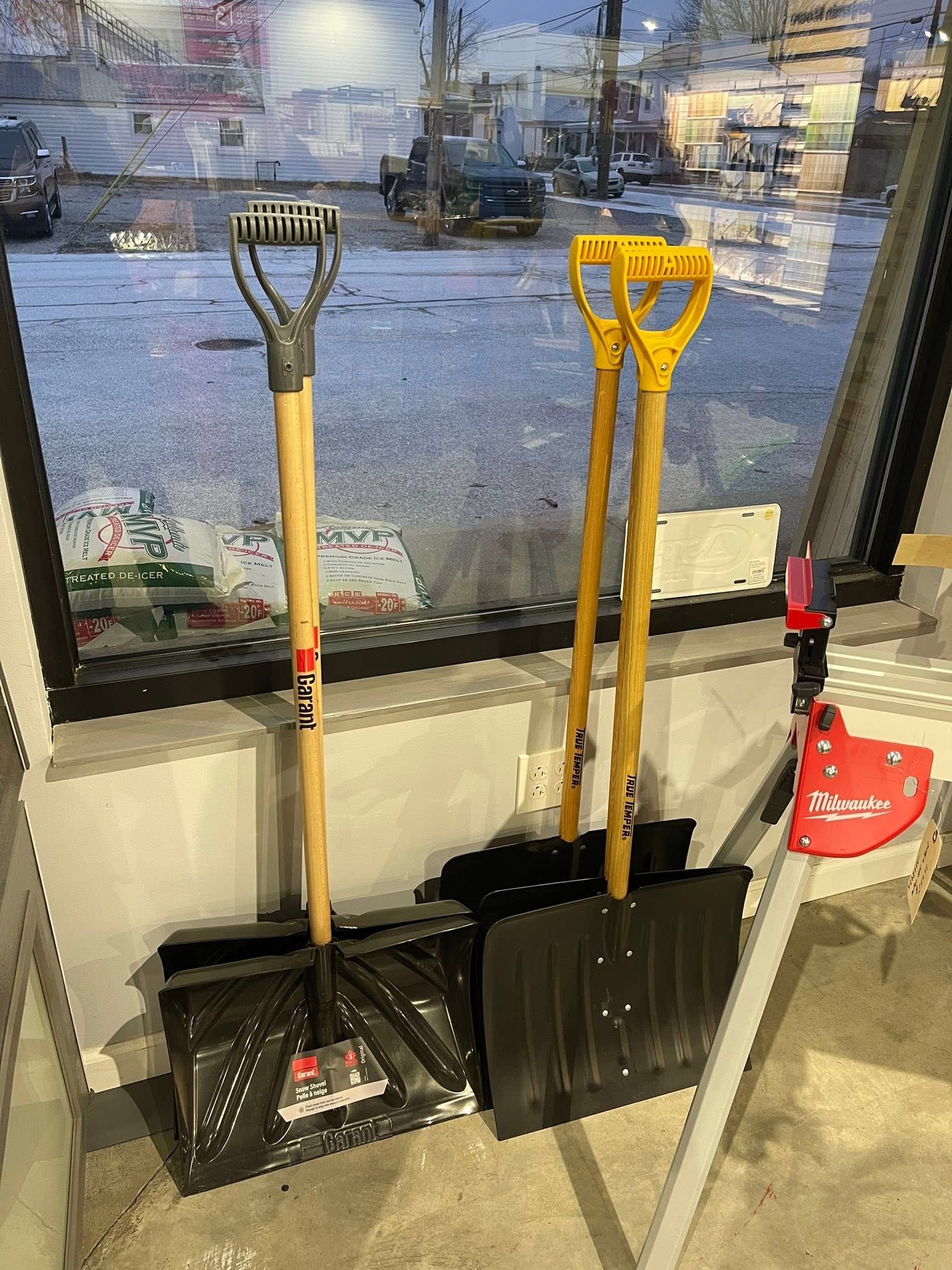 Shovels and a red snow blower by a window, with snowy city buildings outside.
