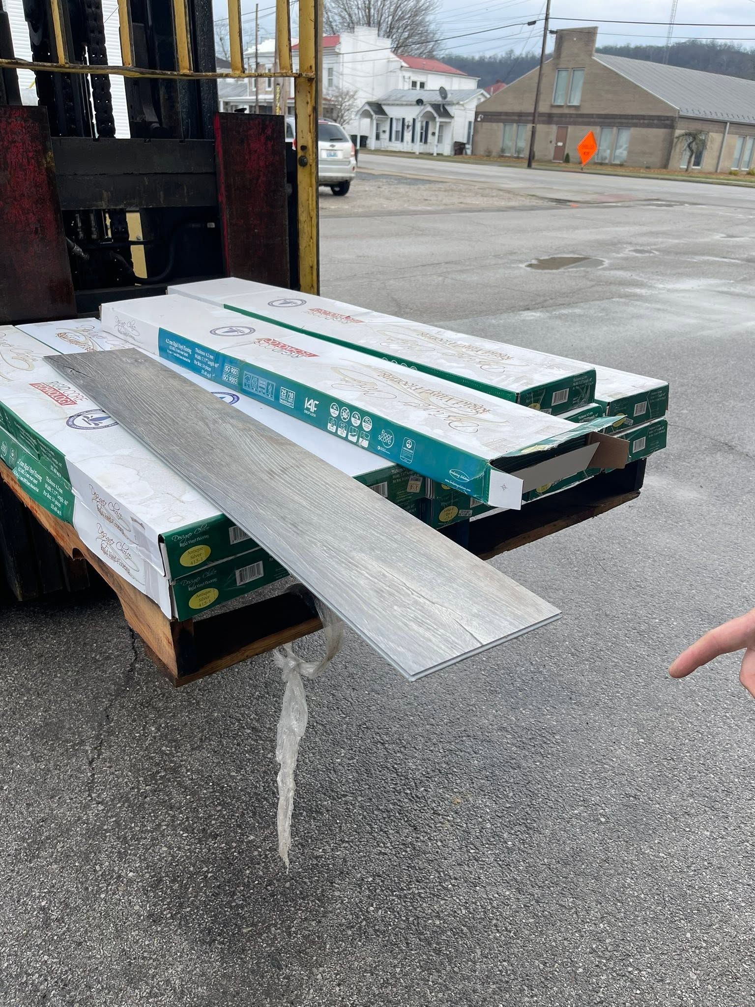 Stacks of long construction boards on a pallet outdoors, with a worker’s hand visible at right.