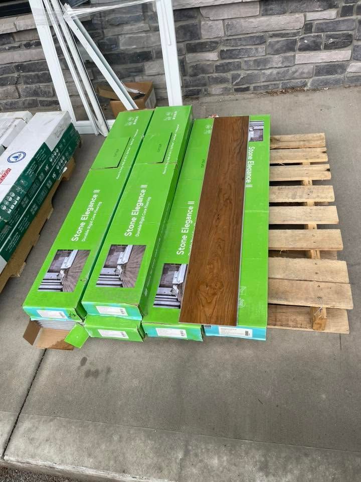 Stacked green and brown flooring boxes on a wooden pallet beside a brick wall