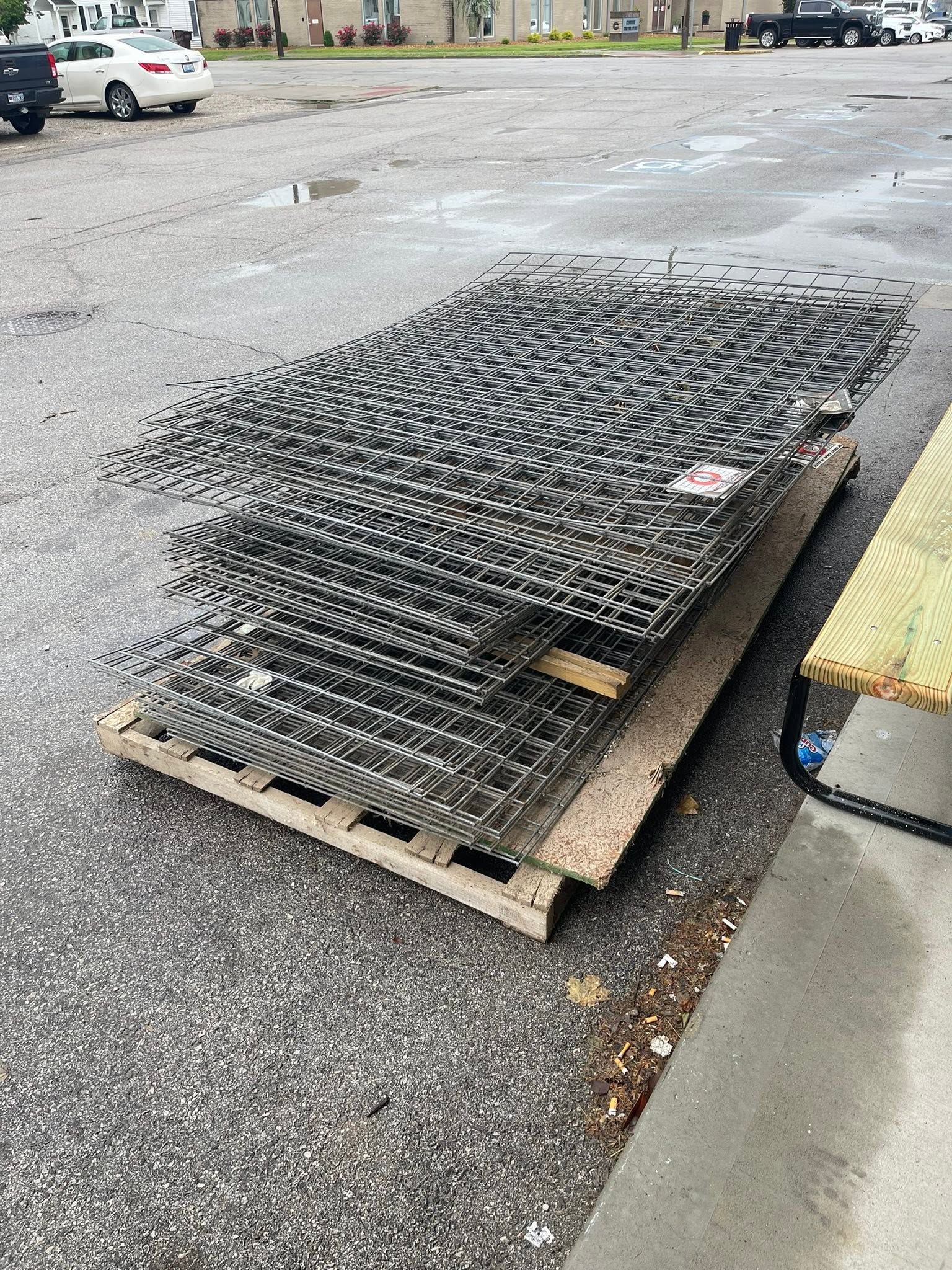 Stacked black metal grates on a wooden pallet beside a curb in a parking lot