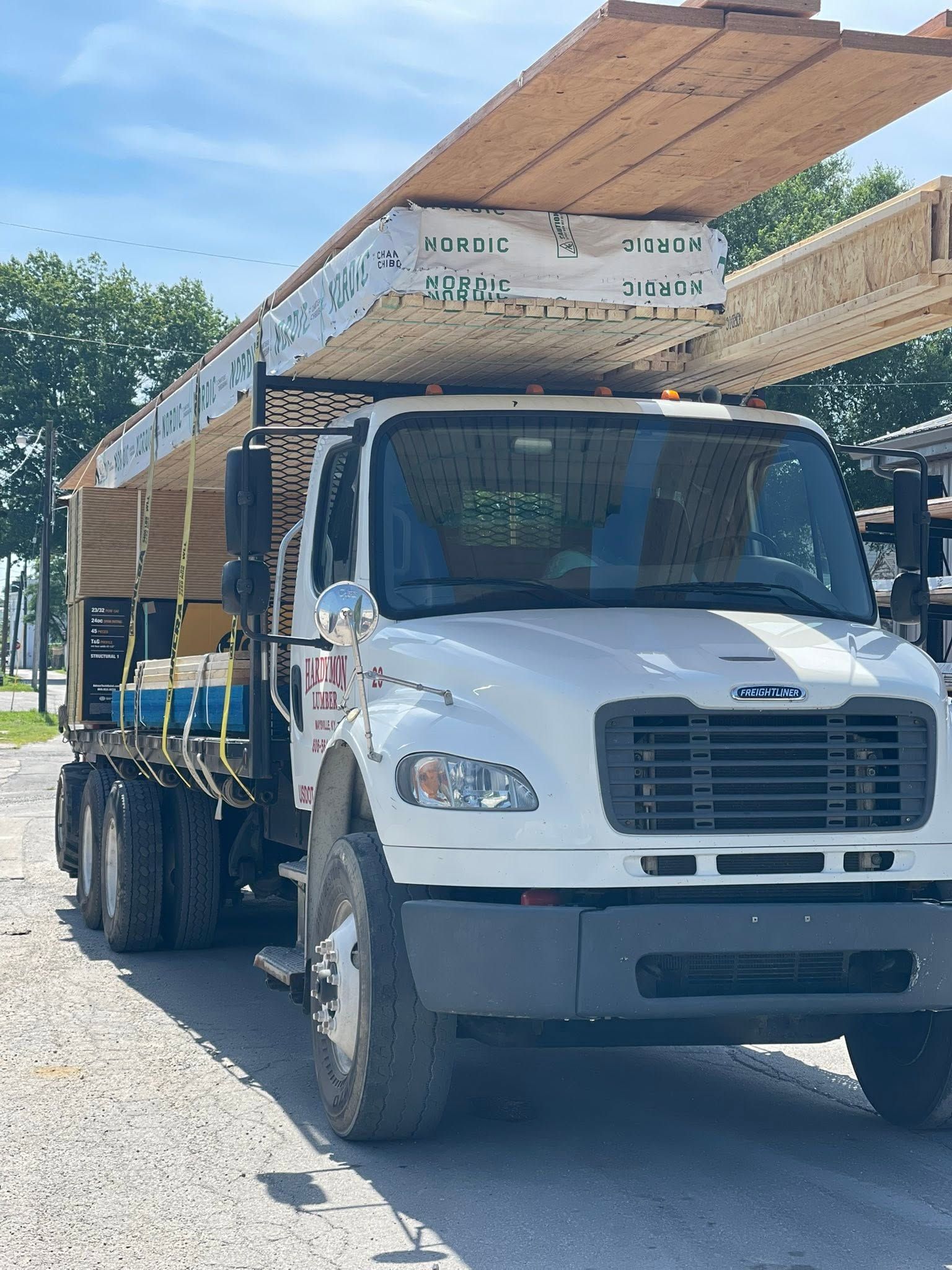 White flatbed truck carrying stacked wood on a sunny street under a canopy of trees