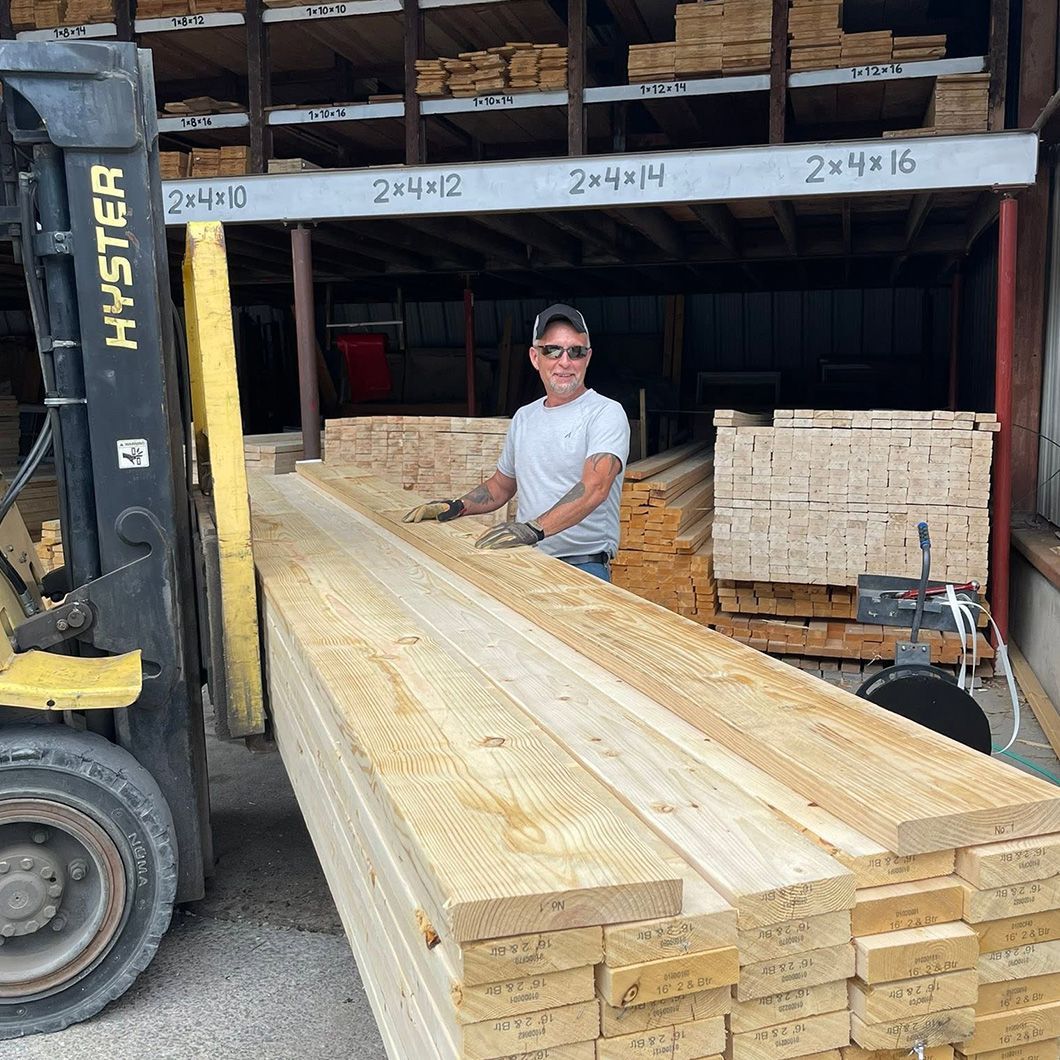 Forklift loading stacked lumber in a warehouse; worker stands beside long wooden boards.