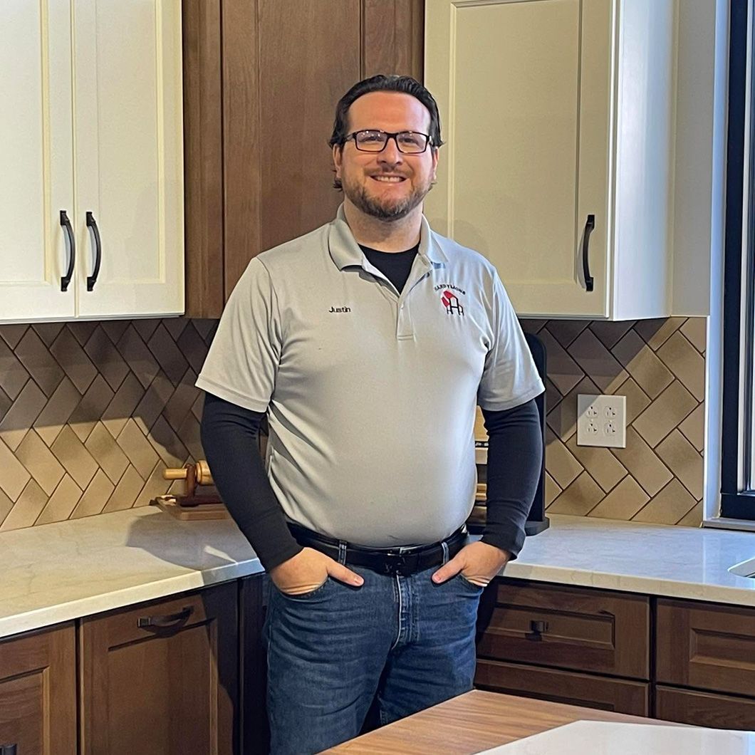 Man standing in a kitchen with hands in pockets, wearing a gray polo and jeans, smiling at the camera.