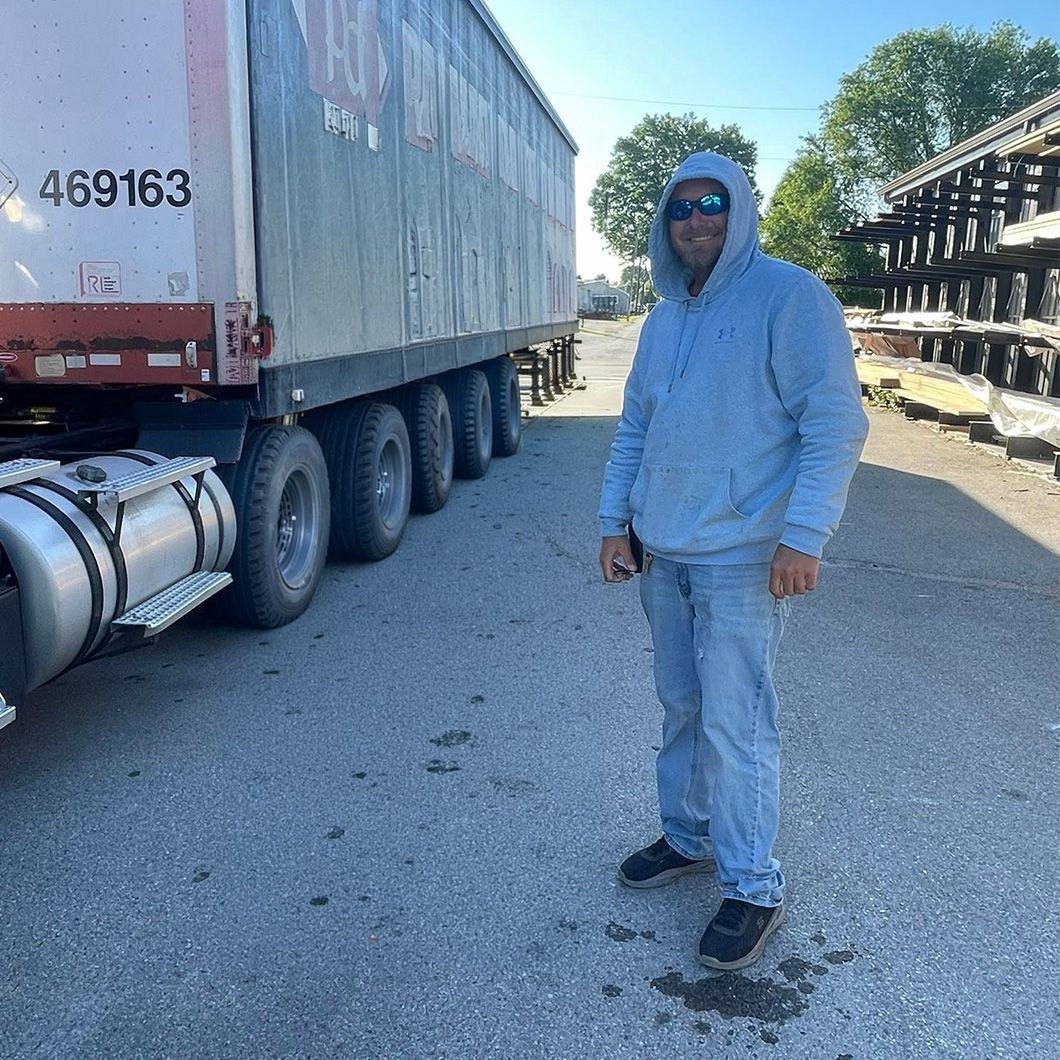 Person in a gray hoodie and jeans stands beside a semi-truck in a gravel lot on a sunny day.
