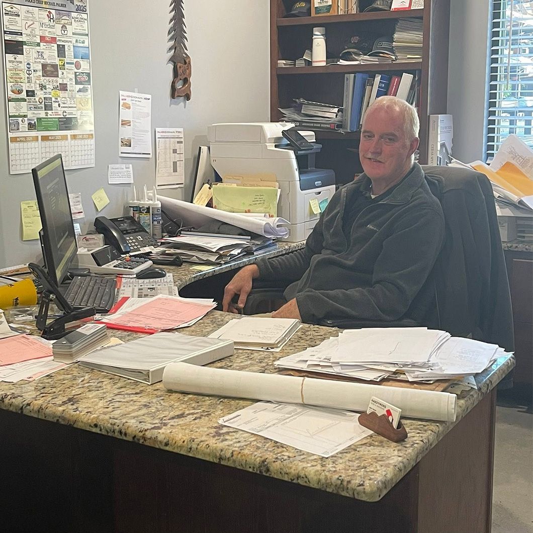 Office worker seated at a cluttered desk, smiling beside a computer and stacks of papers.