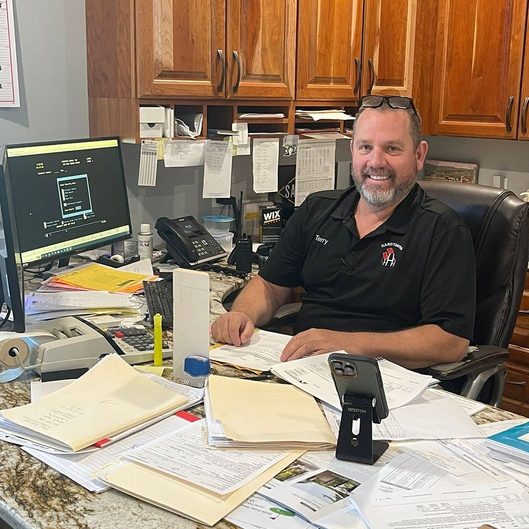 Man smiling at desk in office, surrounded by papers and a computer monitor, wearing a black polo.