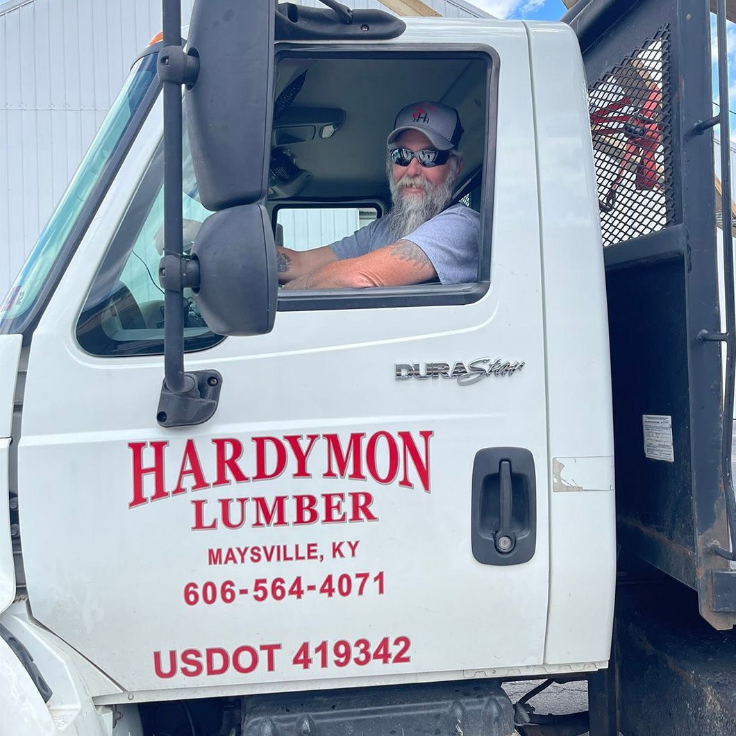White lumber truck cab with HARDY M LUMBER logo and a driver wearing sunglasses inside.