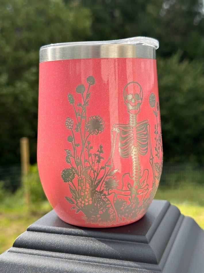 A coral-pink glitter tumbler featuring a laser-engraved skeleton surrounded by wildflowers, set on a post outdoors.