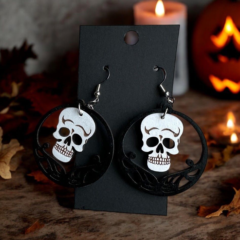 A pair of white skull-shaped earrings inside black circular frames, displayed on a card against a Halloween background.