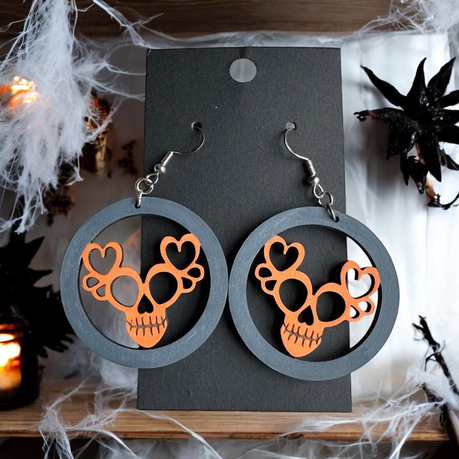 Orange heart-shaped skull earrings in grey circular frames, displayed on a black card against a spooky Halloween backdrop.