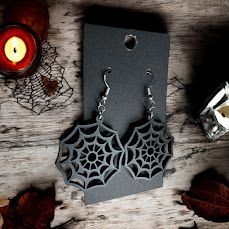 Dark gray spiderweb-shaped earrings on a black display card, set against a rustic wooden background with a red candle.
