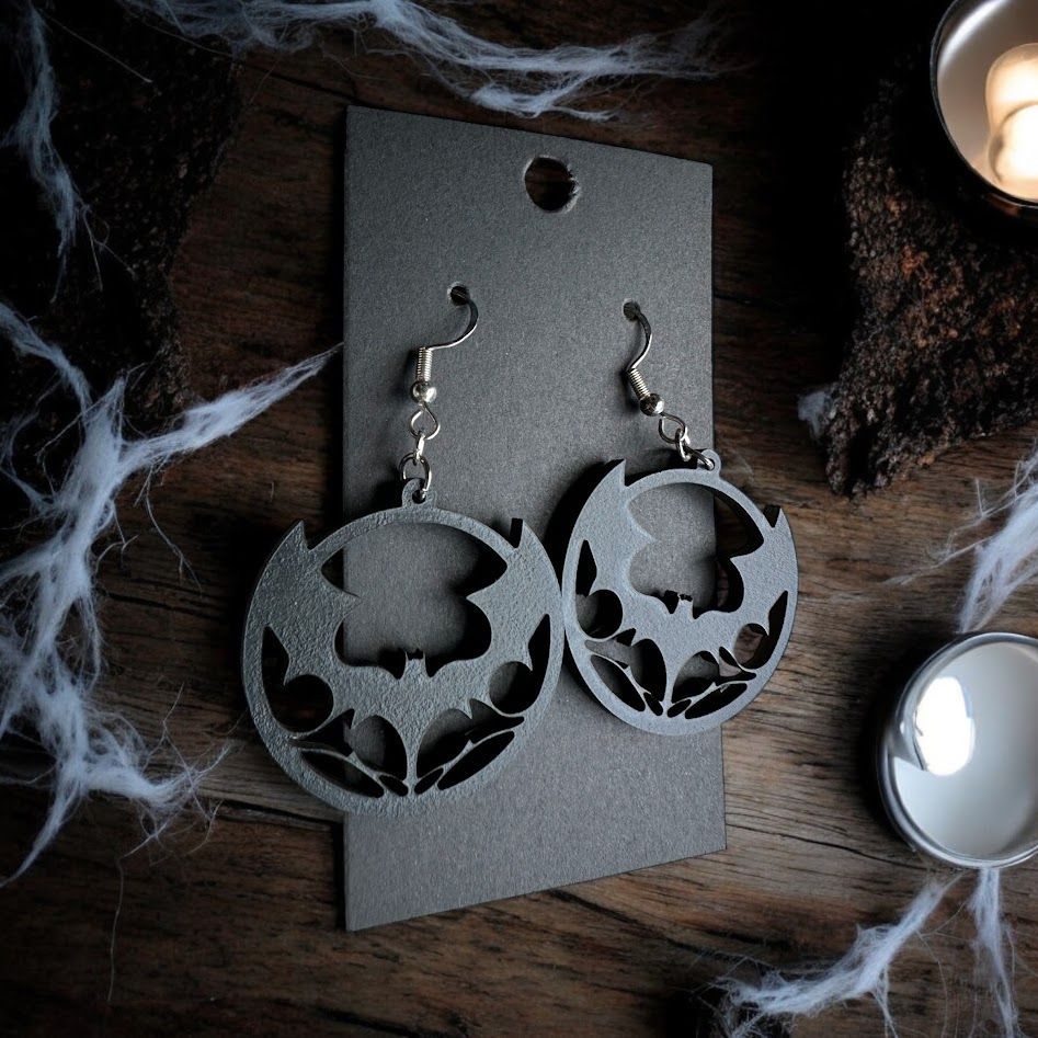 Silver, circular earrings with cutout bat silhouettes, displayed on a grey card against a wooden, cobweb-covered surface.