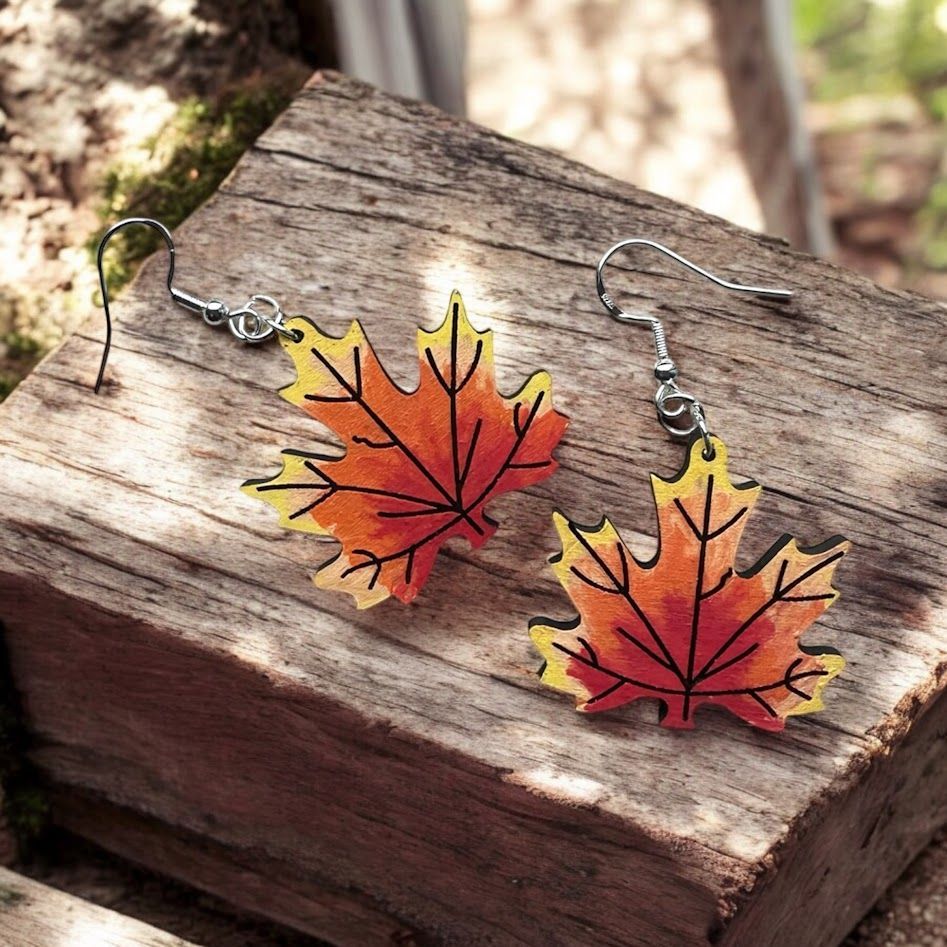 A pair of watercolor maple leaf earrings, featuring gradients of yellow, orange, and red, resting on a rustic wood block.
