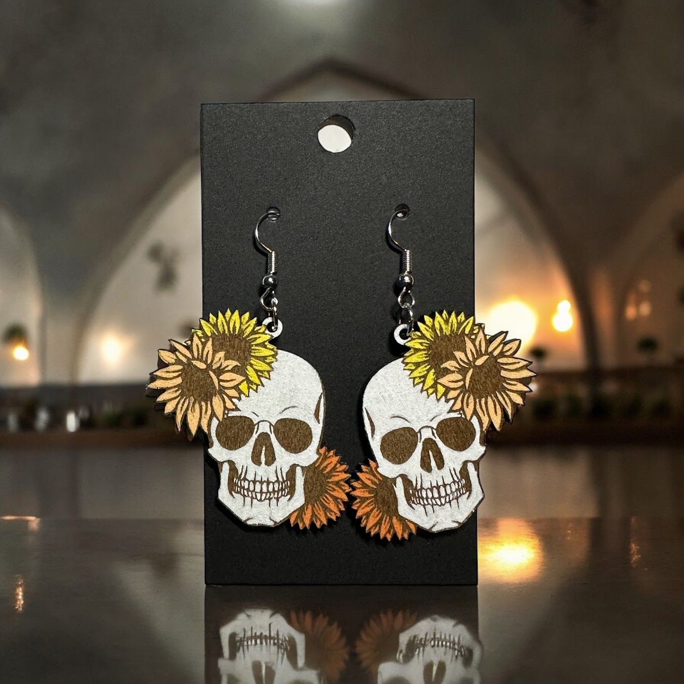 Earrings on a black display card featuring white skulls decorated with yellow sunflowers, hanging from silver hooks.