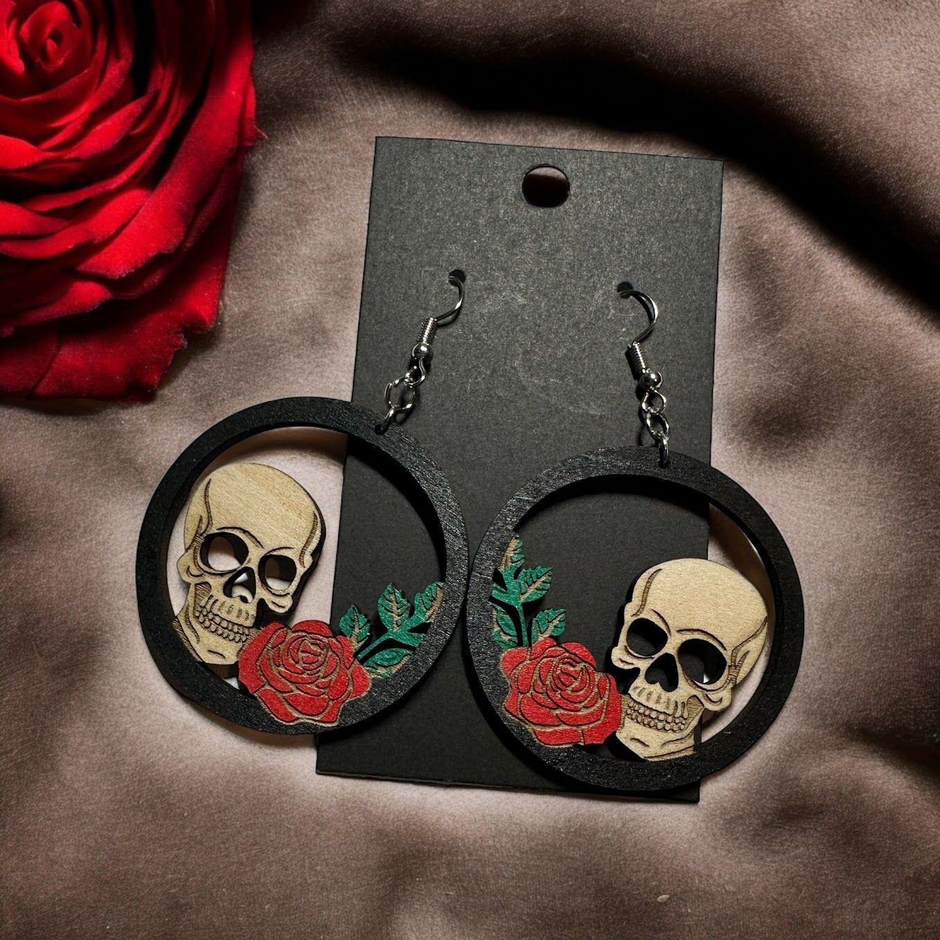 Round black earrings featuring wooden skulls and red roses, displayed on a black card next to a red rose.