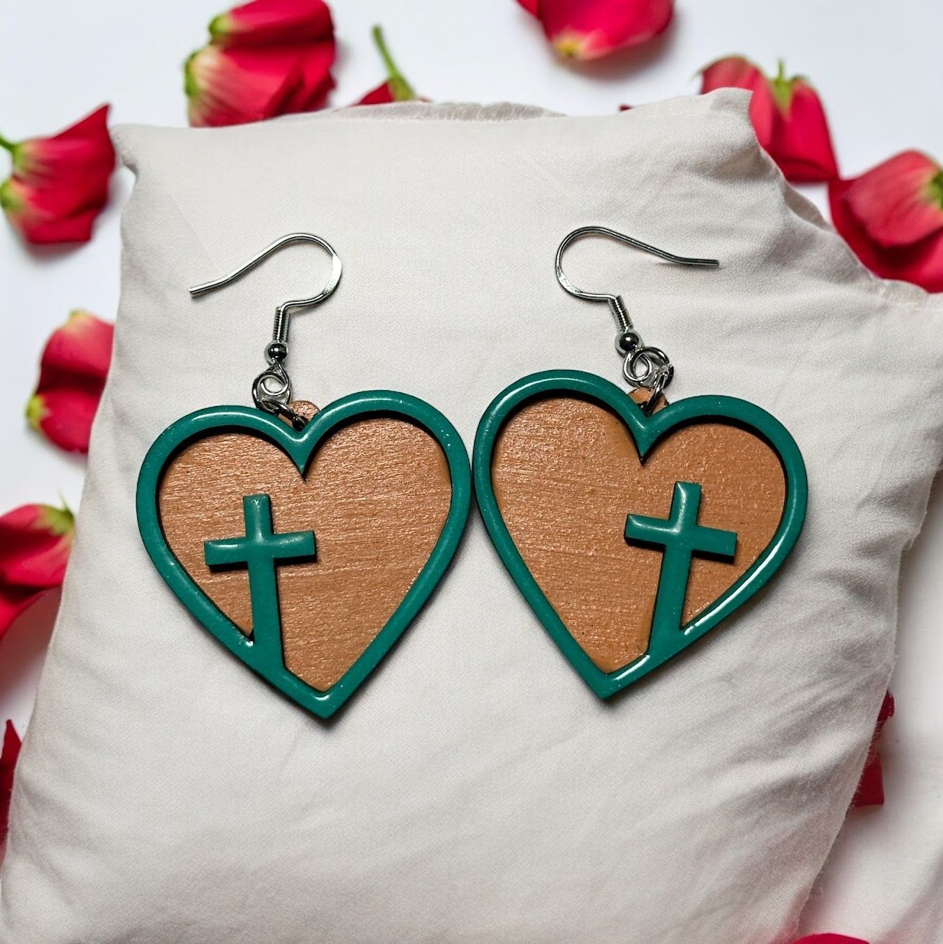 Teal heart-shaped earrings with a tan glitter center and a teal cross overlay, resting on a white cushion with rose petals.