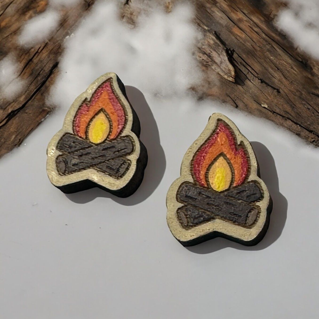 Two wooden, hand-painted campfire earrings with a yellow and orange flame resting on dark brown logs.
