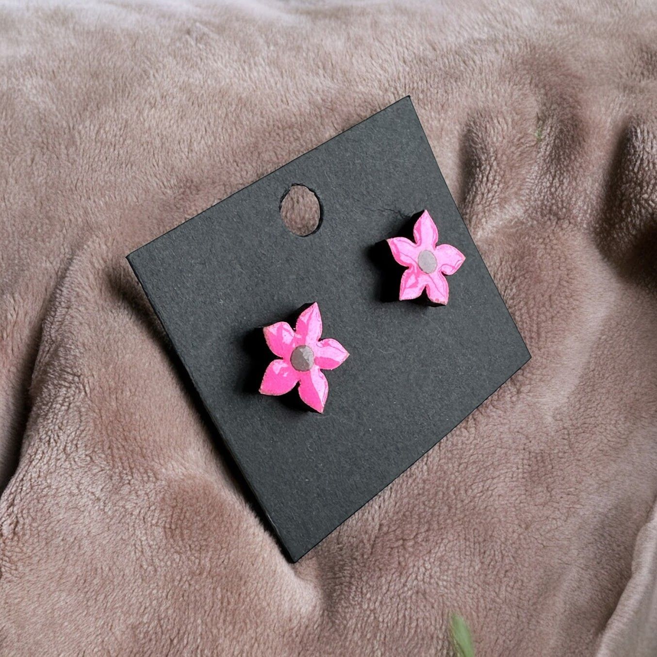 A pair of small, bright pink flower-shaped stud earrings with gray centers, attached to a black display card.