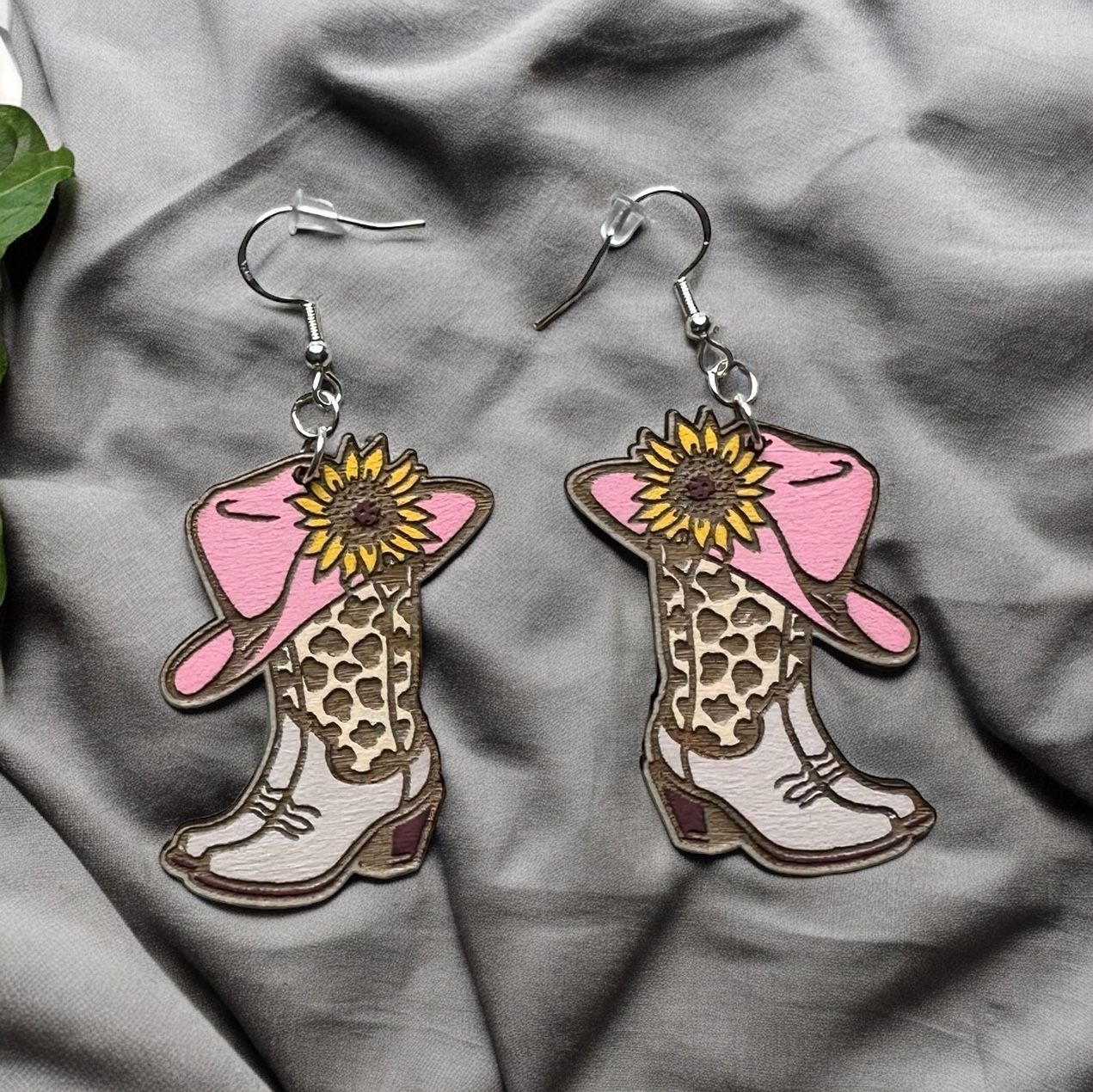 A pair of wooden earrings shaped like cowgirl boots topped with a pink cowboy hat and a yellow sunflower.