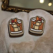 Two wooden, cartoon-style Old Fashioned cocktail pins resting on a plush, cream-colored surface.