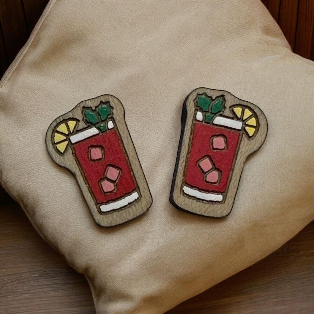 Two wooden drink-shaped ornaments with red liquid, ice cubes, a yellow lemon slice, and green garnish on a beige pillow.