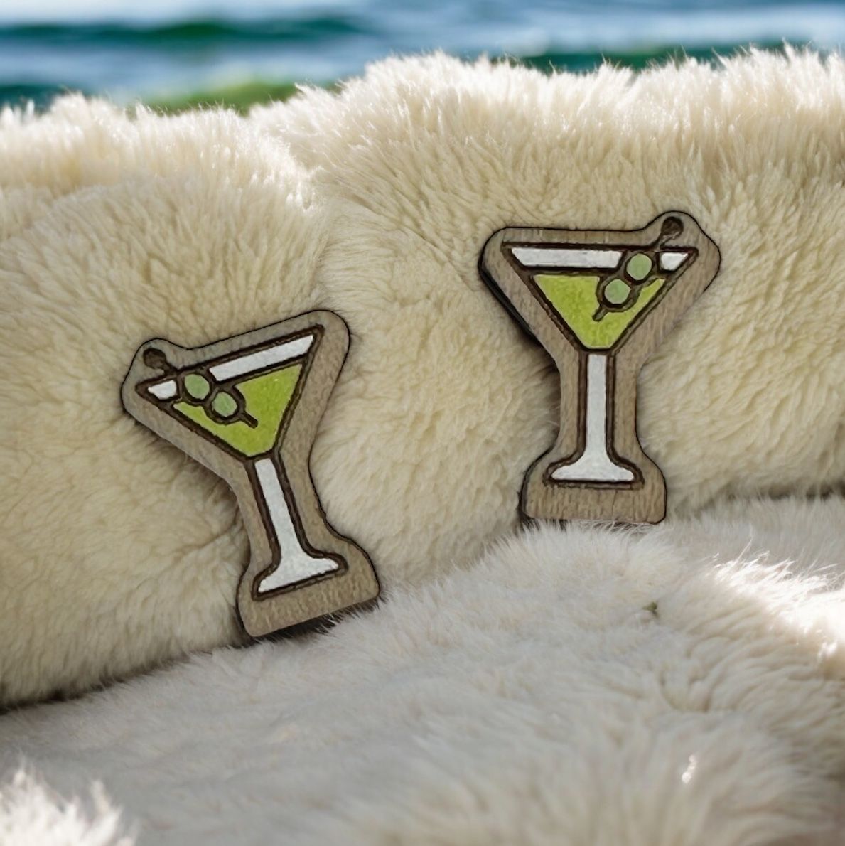Two wooden martini glass ornaments with green liquid and olives resting on fluffy white faux fur with a sea backdrop.