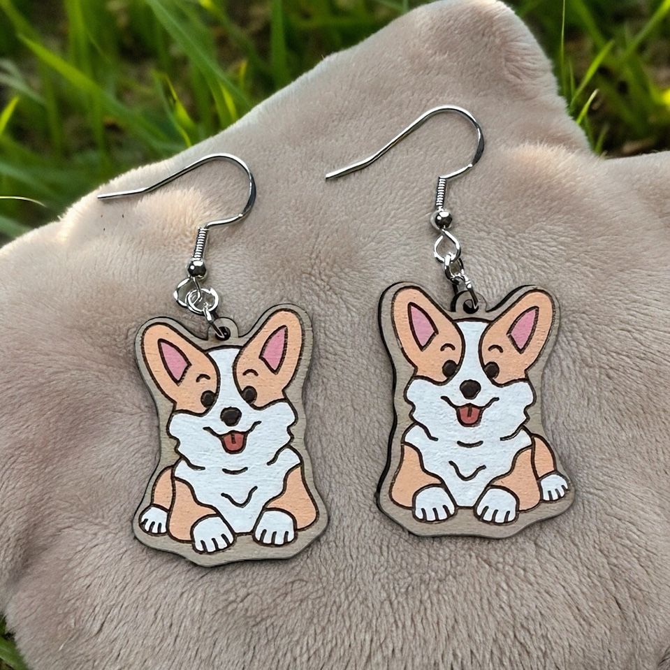 A pair of dangling earrings featuring illustrations of brown and white corgis, set against a fuzzy beige background.
