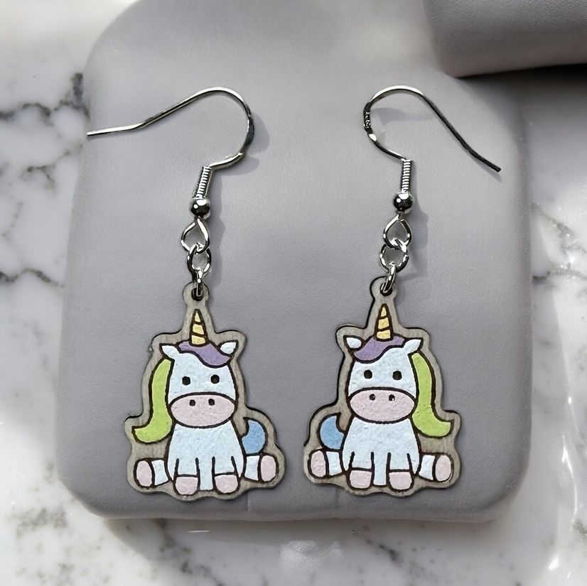 A pair of dangling earrings featuring cute, hand-drawn cartoon unicorns with yellow horns and green manes on a grey stand.