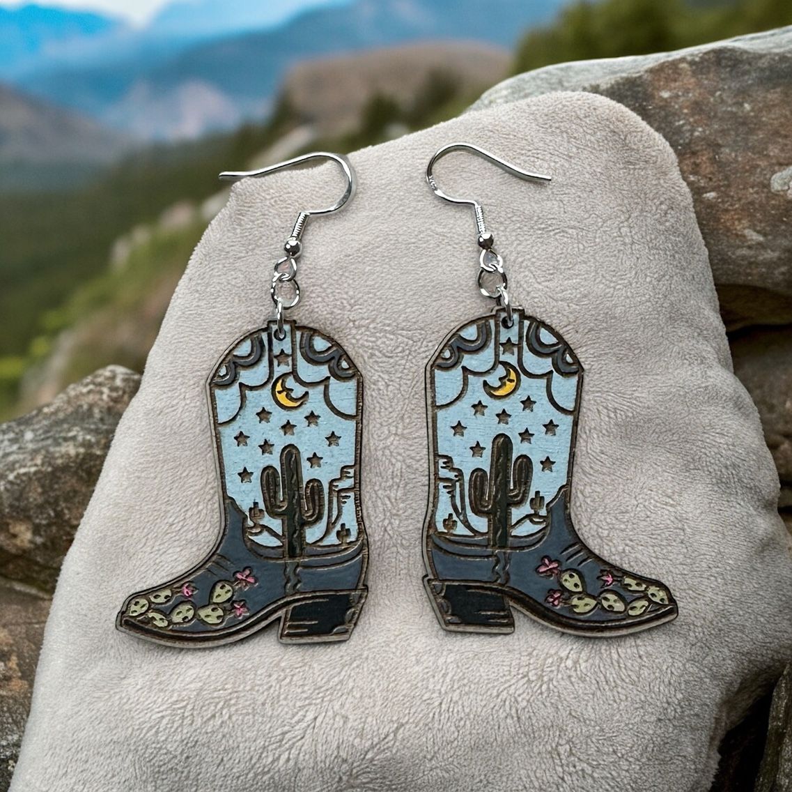 Pair of dangling enamel cowboy boot earrings featuring a desert night scene, displayed on a textured stone surface.