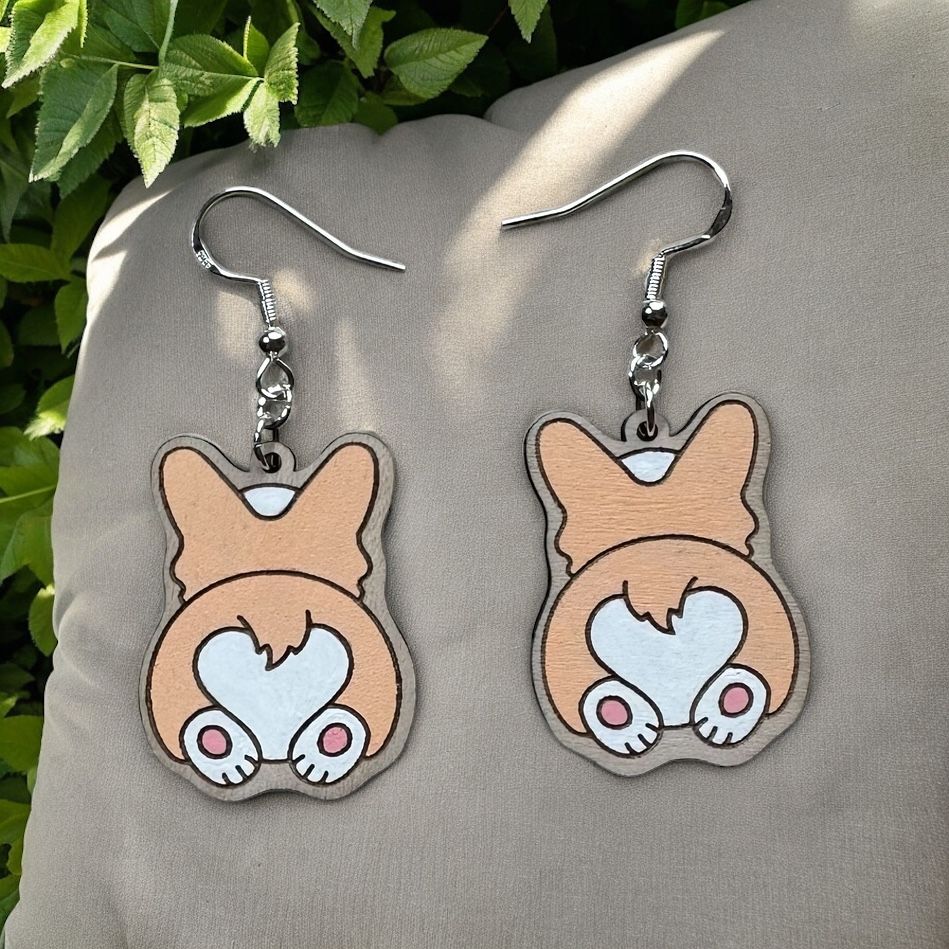 A pair of dangling earrings featuring illustrations of tan and white corgi bottoms with paw pads, resting on a gray pillow.