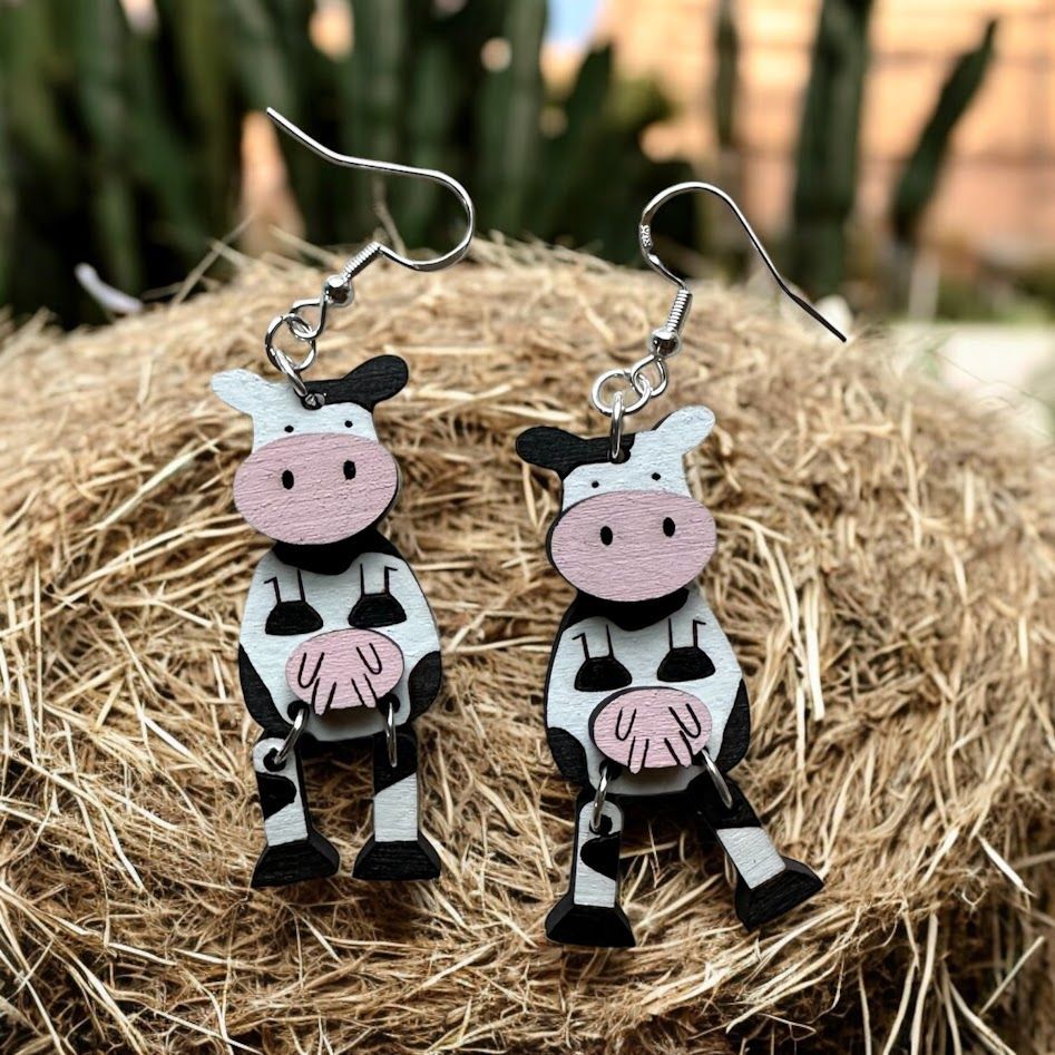 A pair of whimsical cow-shaped wooden earrings with black and white spots and pink muzzles, resting on a bed of hay.