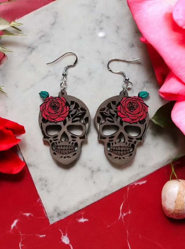 A pair of wood-cut sugar skull earrings with red roses, displayed on a marble surface surrounded by red rose petals.