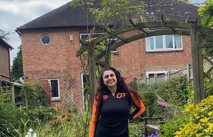 Woman standing in a garden in front of a brick house, smiling under a wooden archway.
