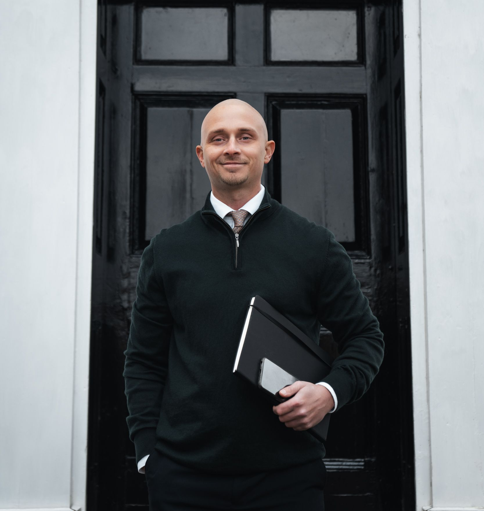 Man standing in a black sweater outside a doorway, holding a folder and smiling