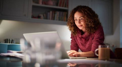Woman working on a laptop at a desk in a home office, wearing a red sweater and smiling.
