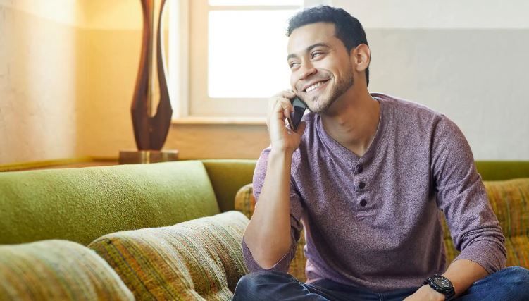 Man smiling while talking on a phone, seated on a green couch in a bright living room.