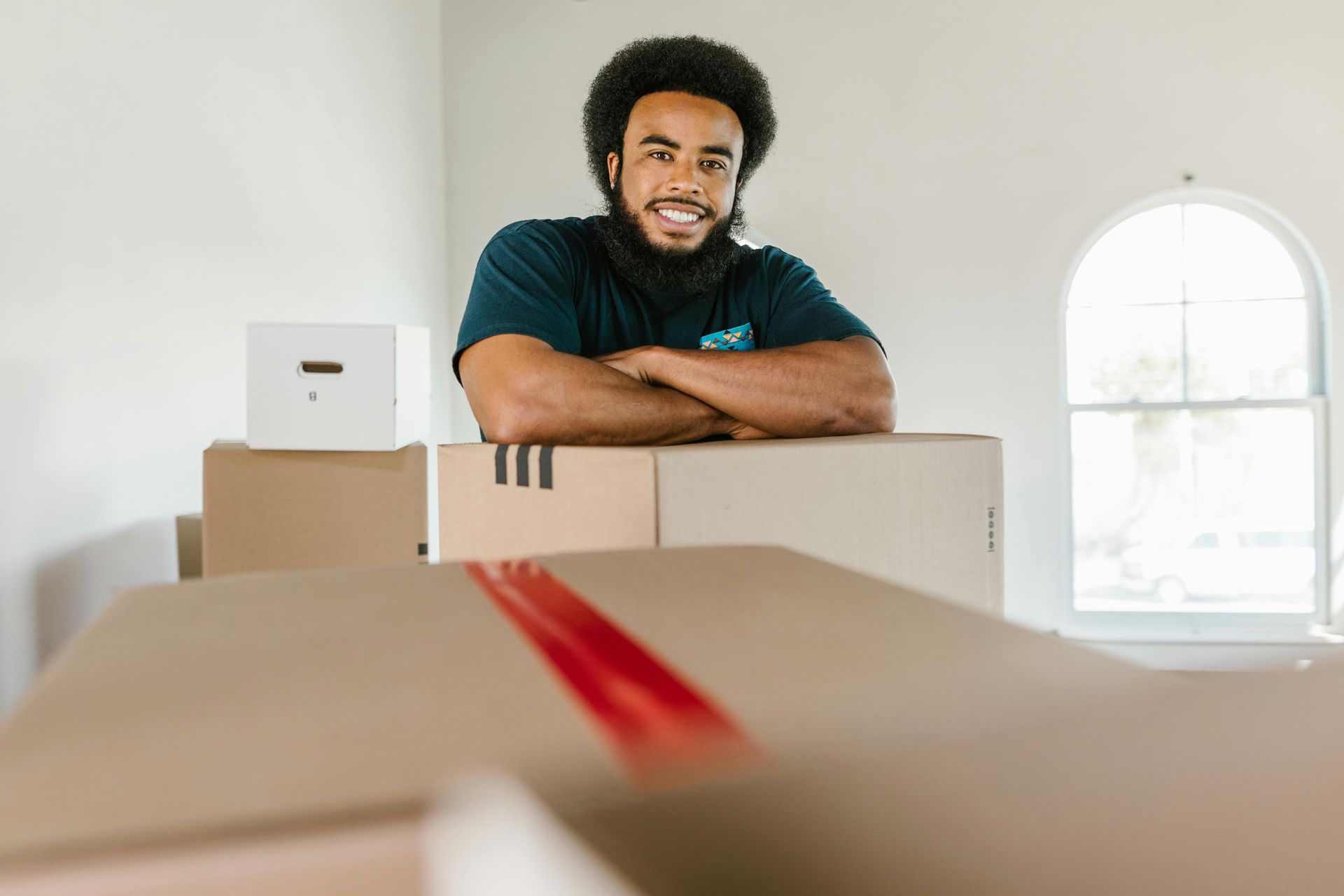 Man smiling behind stacked moving boxes in a bright room, with a laptop on one box