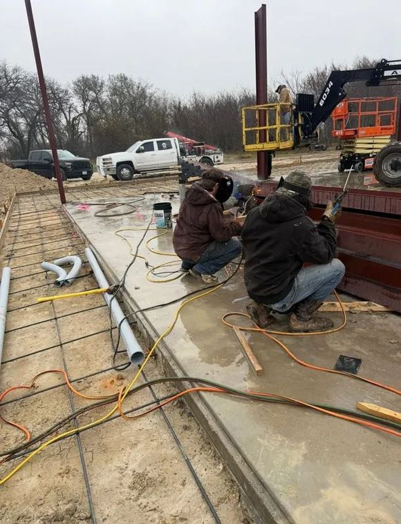 Workers welding steel beams on a concrete site beside a crane and parked trucks.