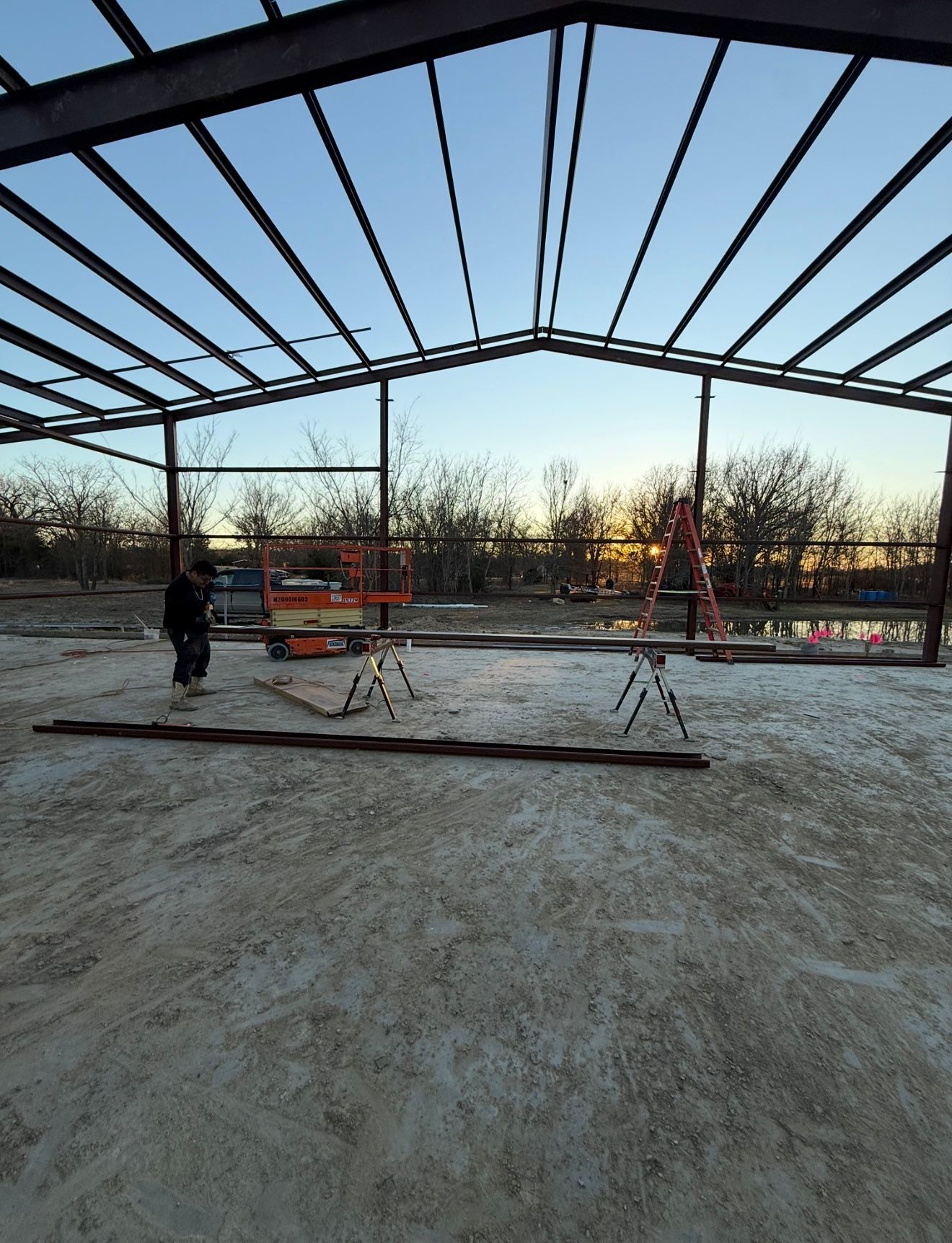 Steel building frame under construction at sunset, with workers, ladders, and materials on a dirt floor.
