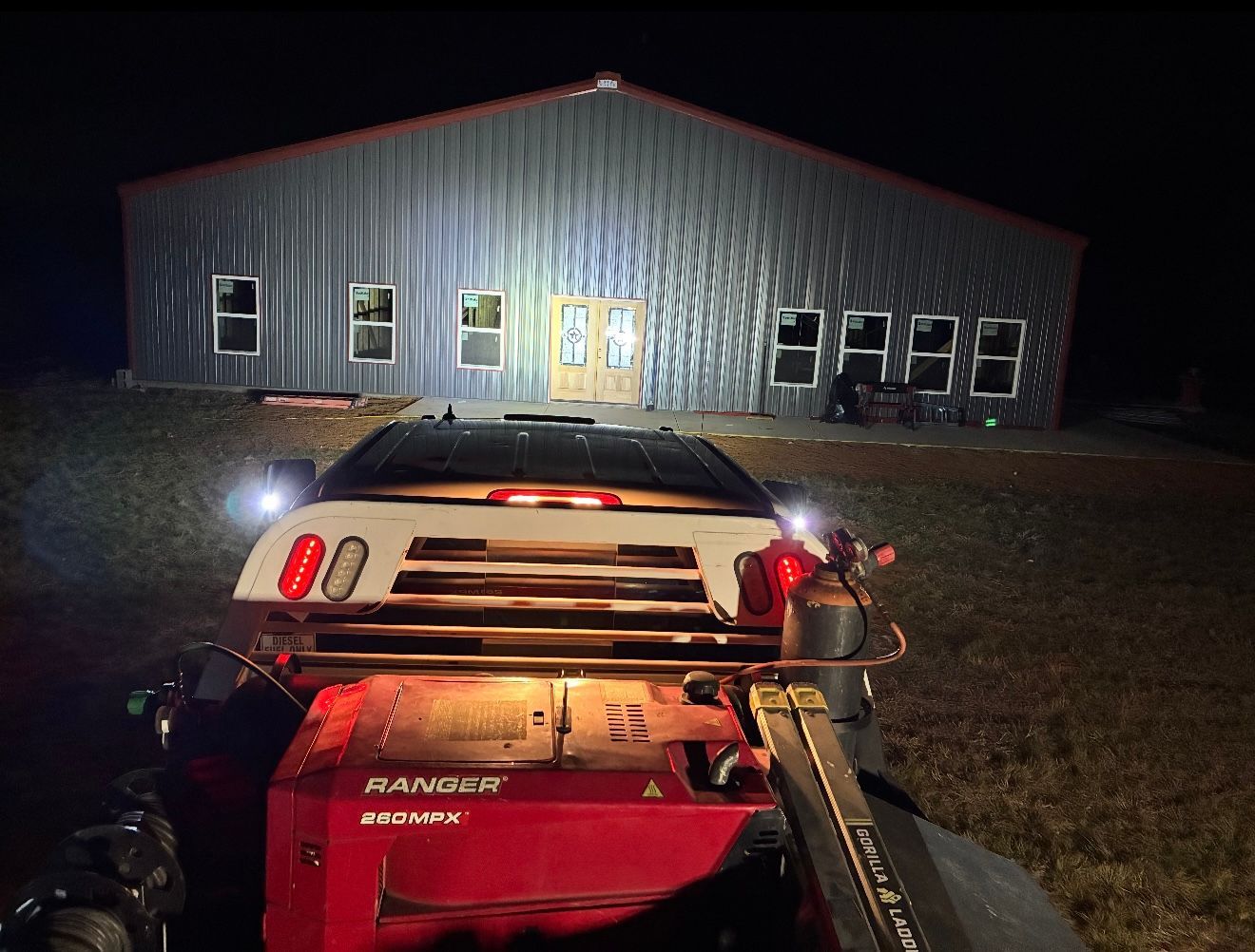 Nighttime view from an ATV toward a lit metal building with several windows and a central door.