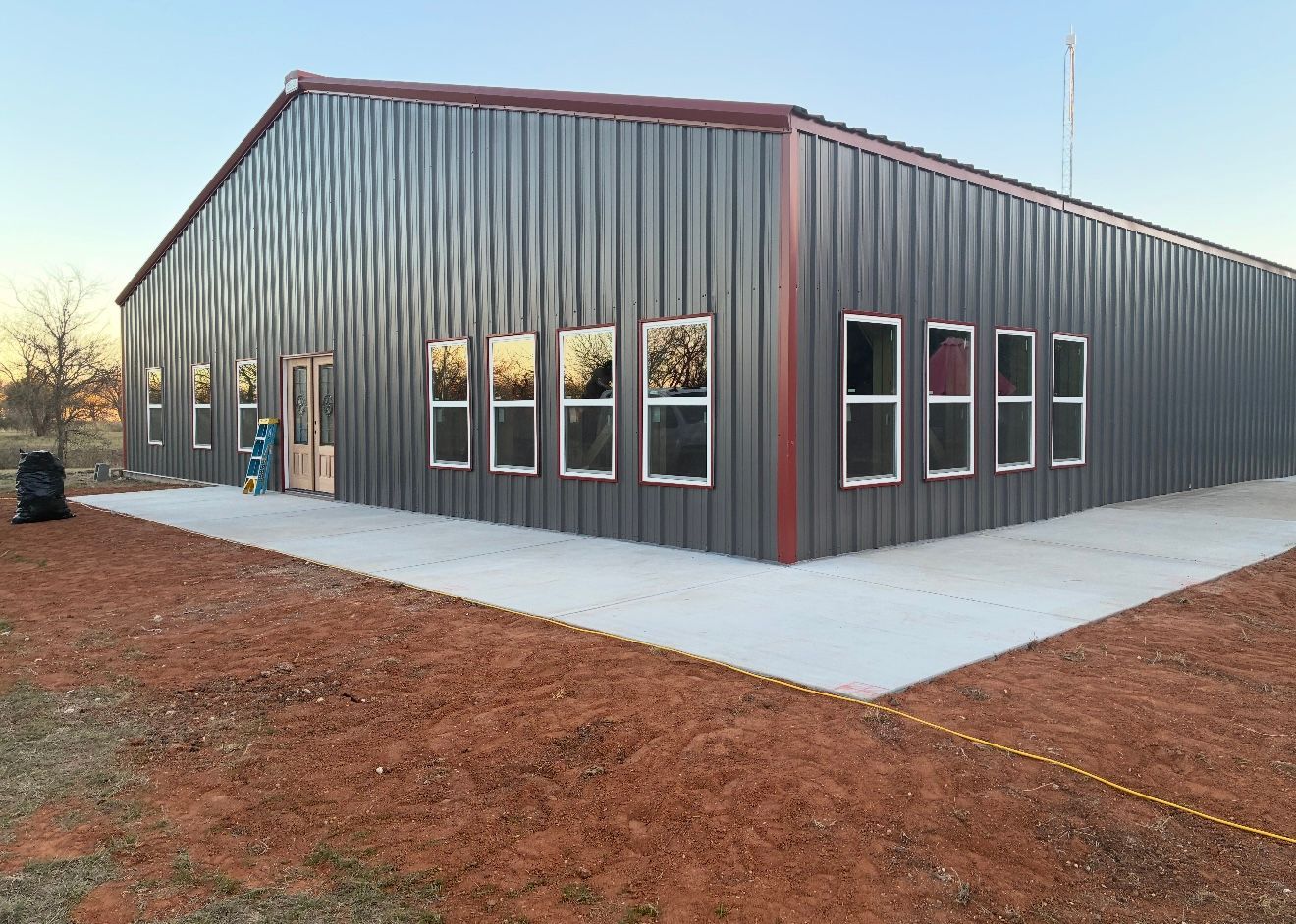 Gray metal warehouse building with red trim, multiple windows, and a concrete apron on a dirt lot