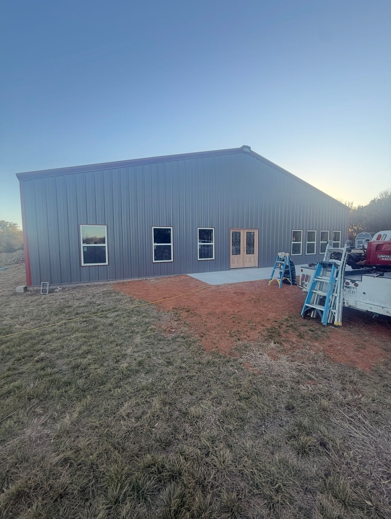 Gray metal building at dusk with several windows, grassy yard, and construction equipment in front