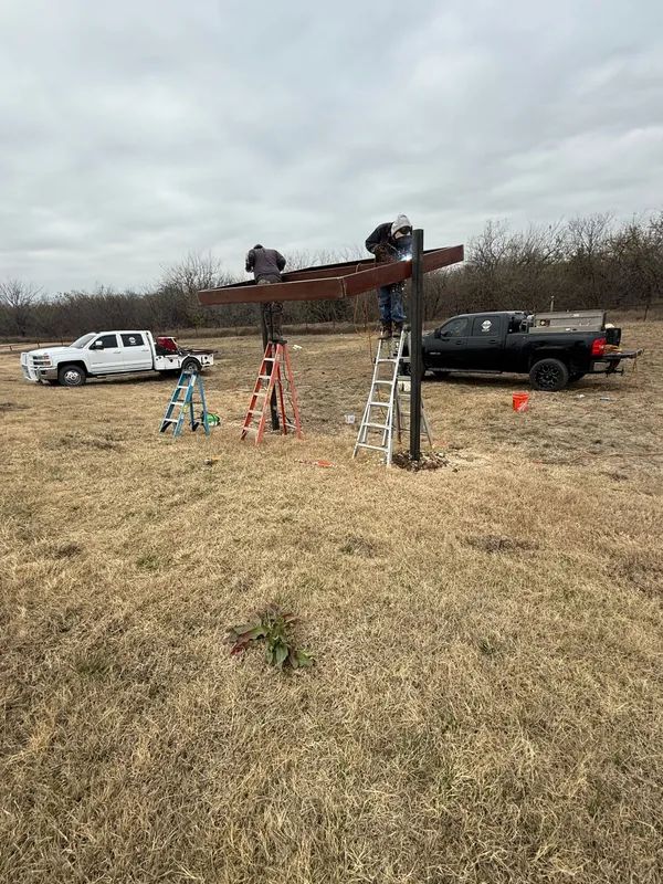 Workers atop a large wooden beam between ladders in a grassy field, with trucks parked nearby under cloudy skies