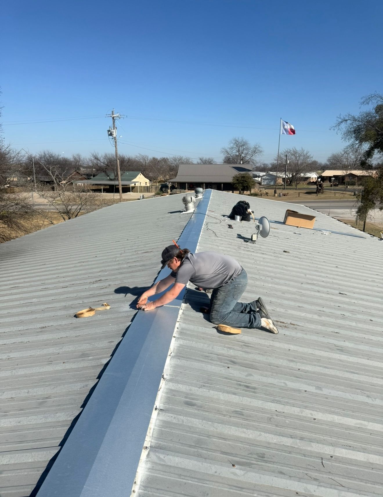 Worker crouching on a metal roof, securing a long blue seam under a clear sky.