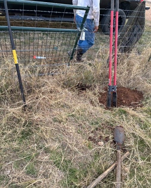 Red fence posts being driven into a grassy field near a gate and shovel, with a person partially visible.